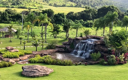 Cachoeira pequena em meio a gramado plano com árvores ao redor e construção ao fundo