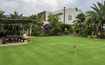 Campo de grama verde bem nivelado em jardim residencial com palmeiras, pergolado de madeira, mesa externa e casa moderna ao fundo sob céu nublado.