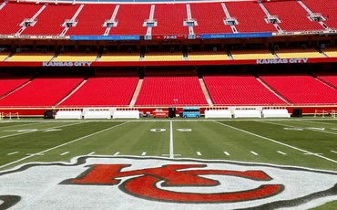 Campo do Arrowhead Stadium, casa do Kansas City Chiefs, com logo da equipe pintado no gramado, visto de um ângulo central com assentos vermelhos ao fundo.