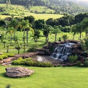 Cachoeira pequena em meio a gramado plano com árvores ao redor e construção ao fundo