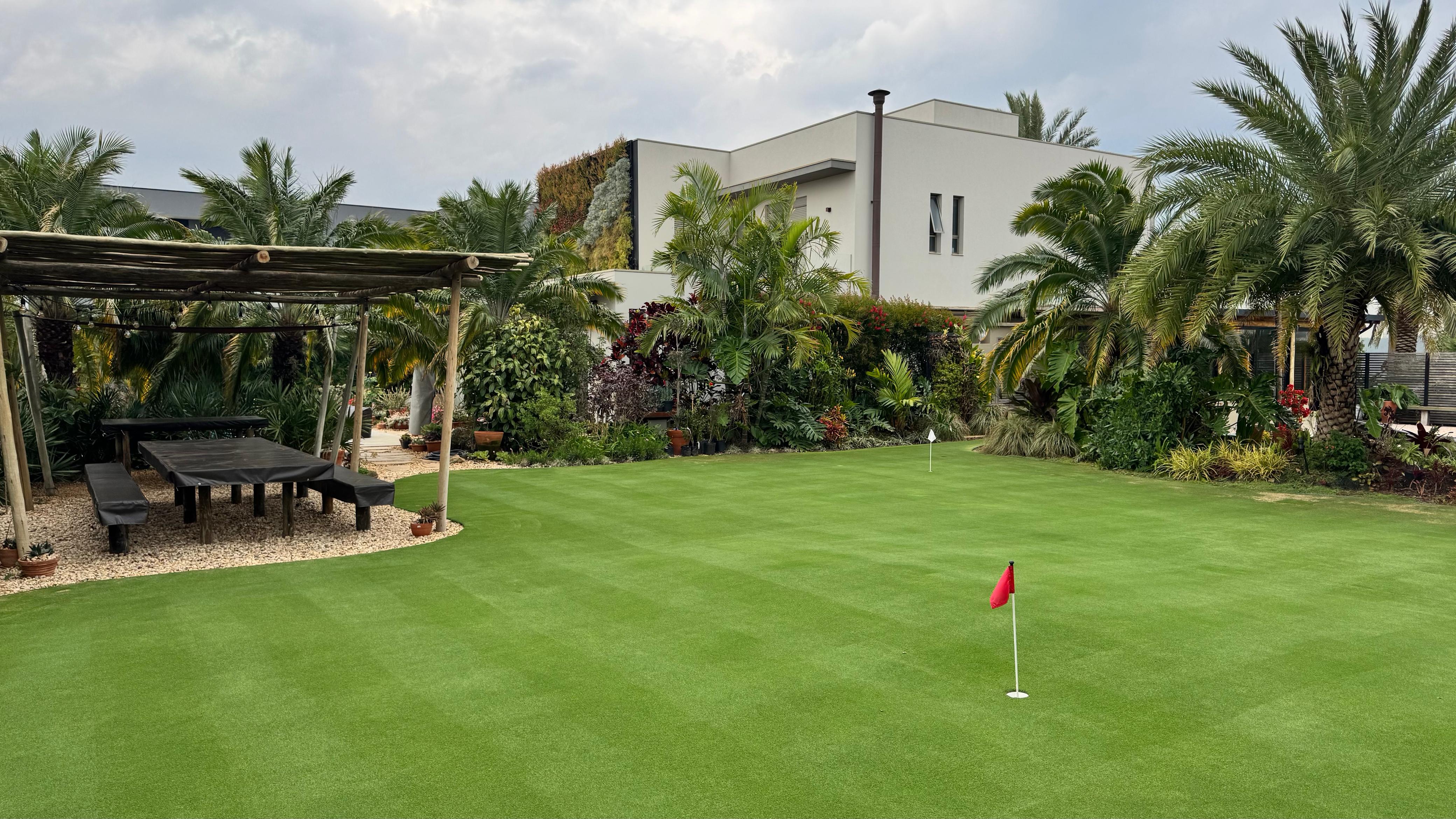 Campo de grama verde bem nivelado em jardim residencial com palmeiras, pergolado de madeira, mesa externa e casa moderna ao fundo sob céu nublado.